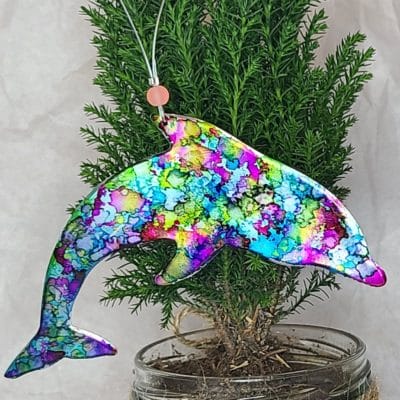 Close-up picture of dolphin-shaped aluminum gift tag/ornament that has been painted with brightly colored acrylic paints in hues of blues, purples and greens. Ornament is hanging on a small cypress tree planted in a glass jar.